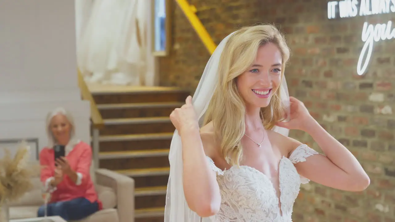 Grandmother Taking Photo Of Adult Granddaughter Choosing And Trying On Wedding Dress In Bridal Store