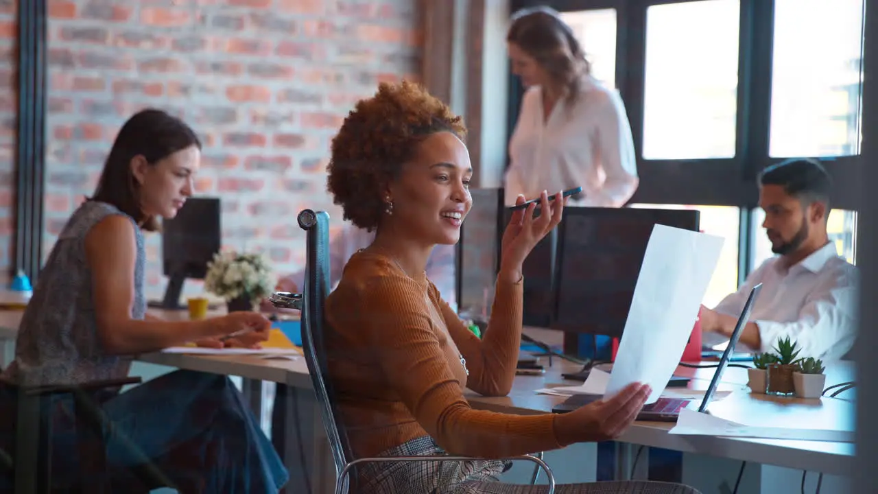 Businesswoman In Office Working On Laptop At Desk Talking Into Mic Of Mobile Phone