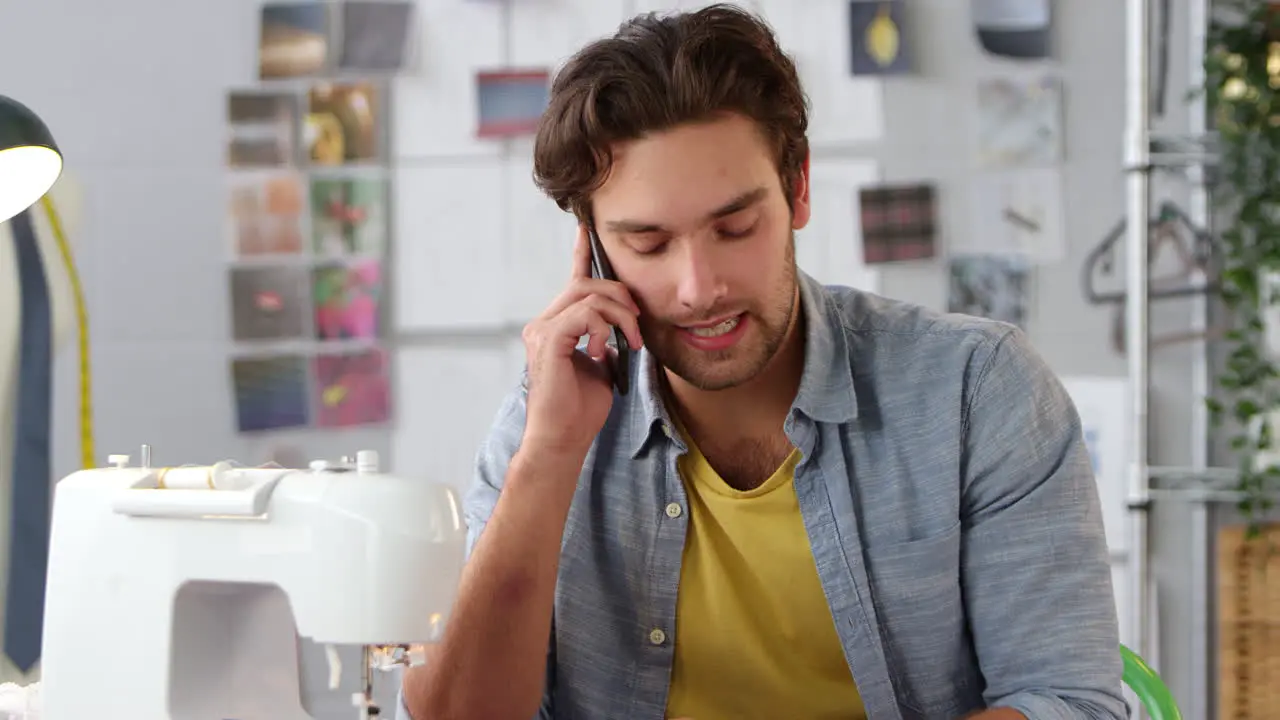 Male Owner Of Fashion Business Making Call On Mobile Phone At Sewing Machine In Studio