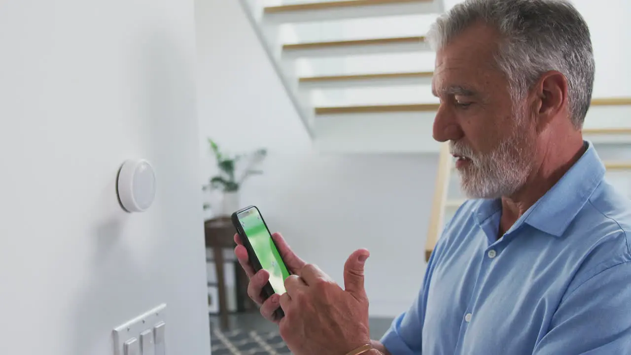 Senior Hispanic Man Using App On Phone To Control Digital Central Heating Thermostat At Home