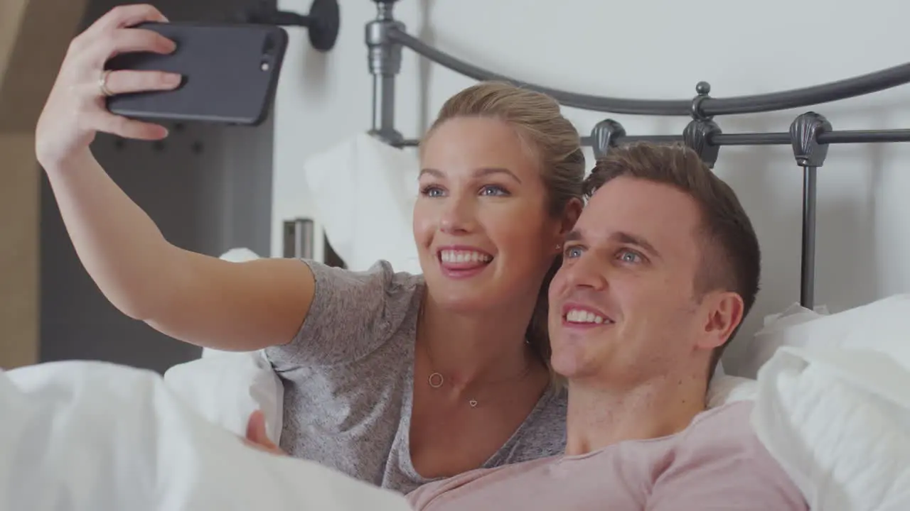 Couple In Bed Wearing Pyjamas Posing For Selfie On Mobile Phone