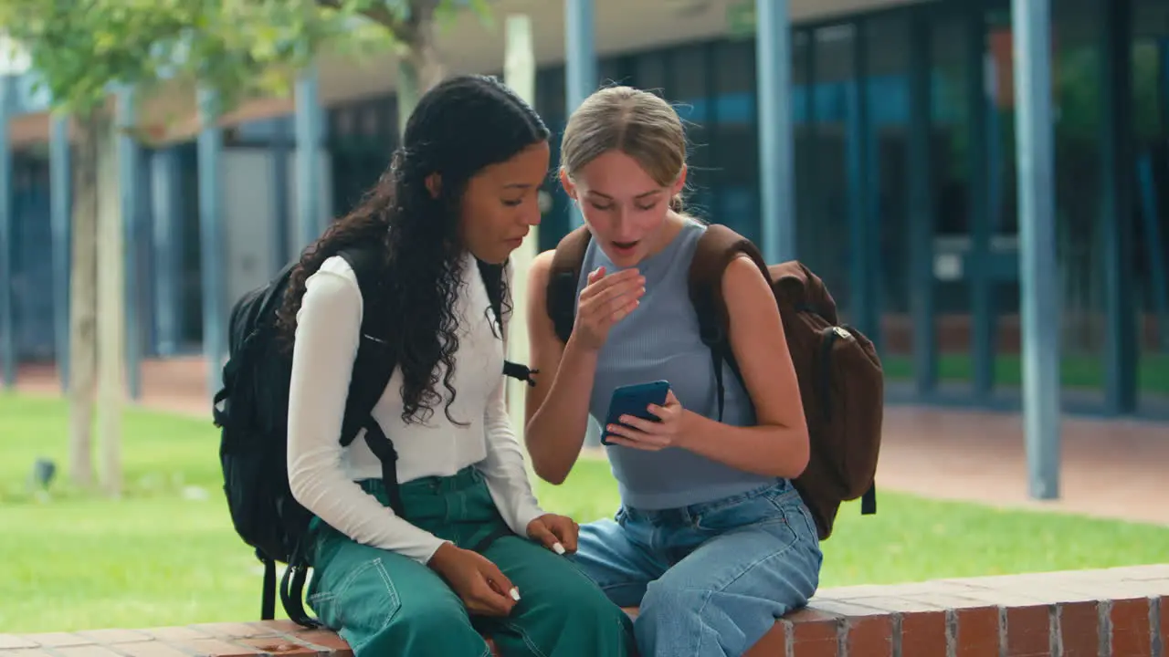 Two Female High School Or Secondary Students Looking At Social Media On Phone Sitting Outdoors