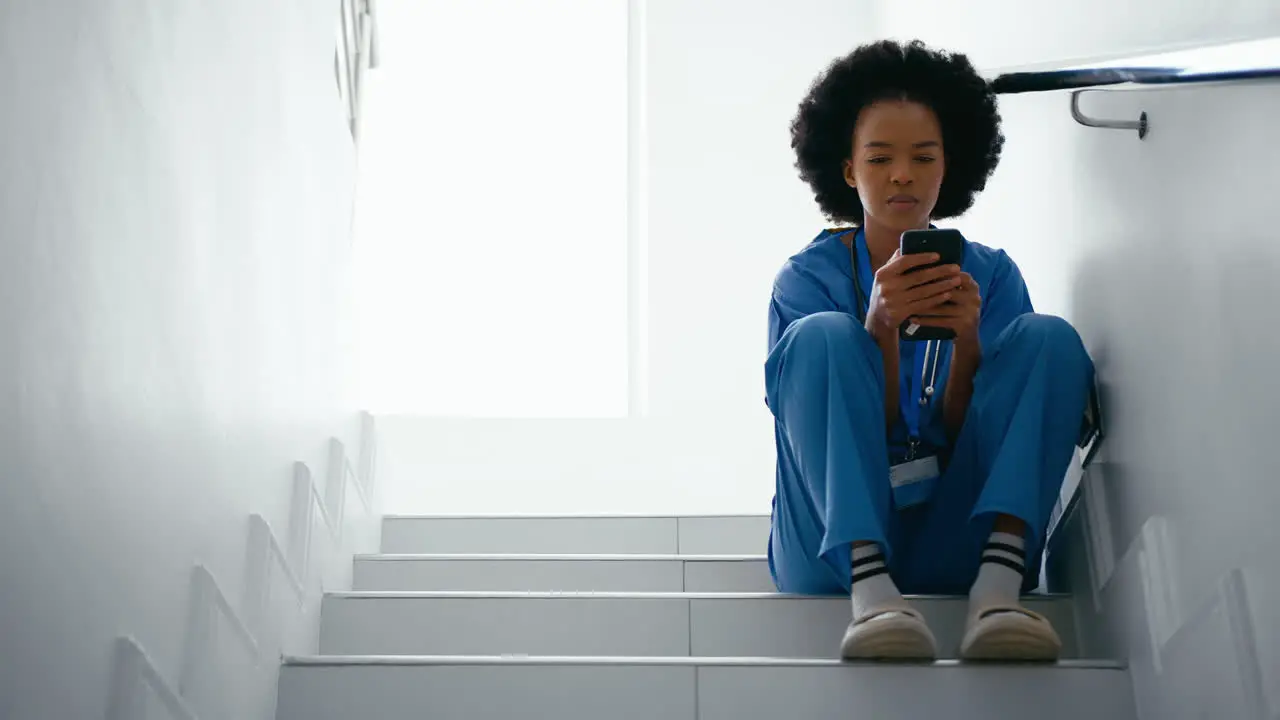 Female Nurse Wearing Scrubs Browsing On Mobile Phone Sitting On Stairs In Hospital