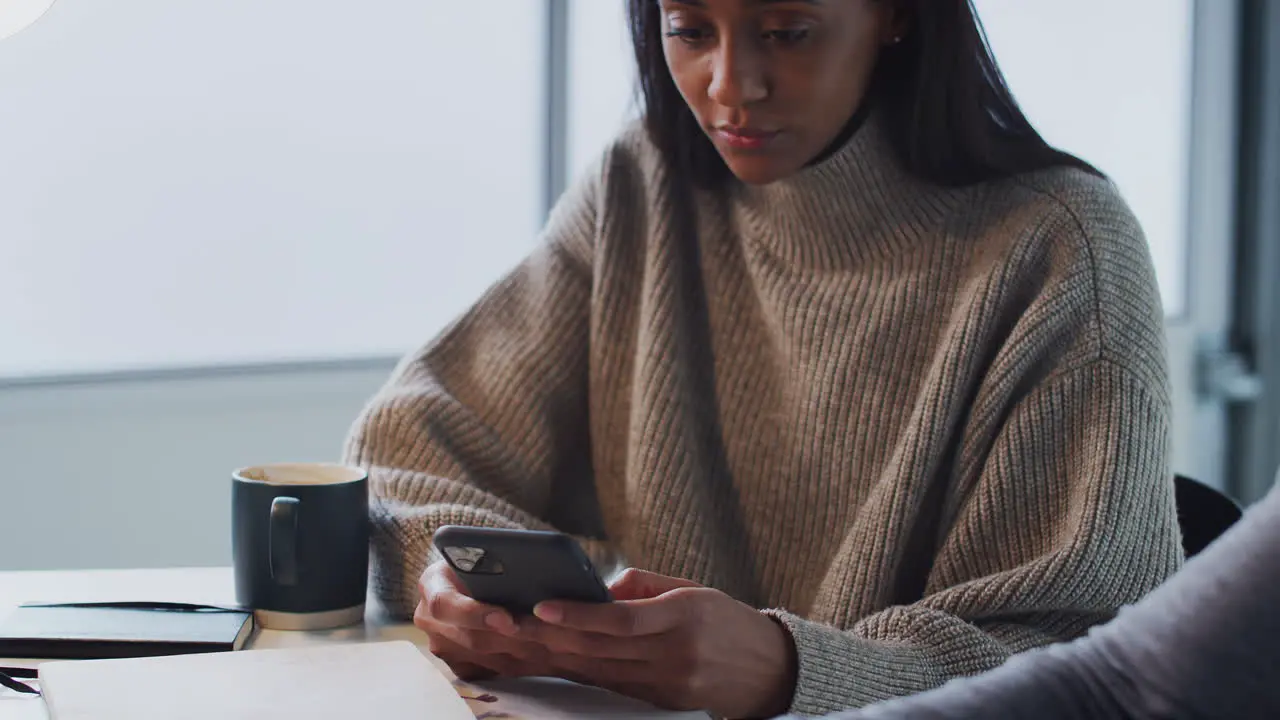 Businesswoman Using Mobile Phone As Male Colleague Works On Laptop In Foreground