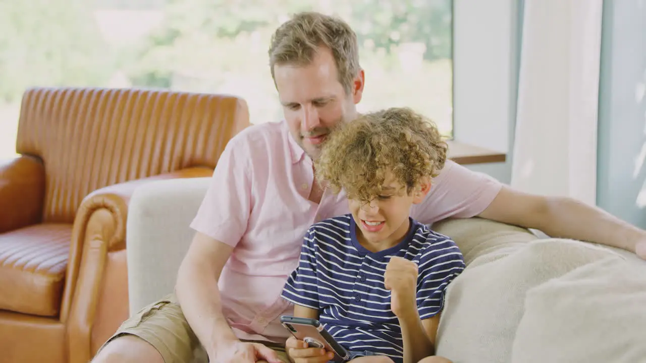 Father And Son Sitting On Sofa At Home Playing Video Game On Mobile Phone Together