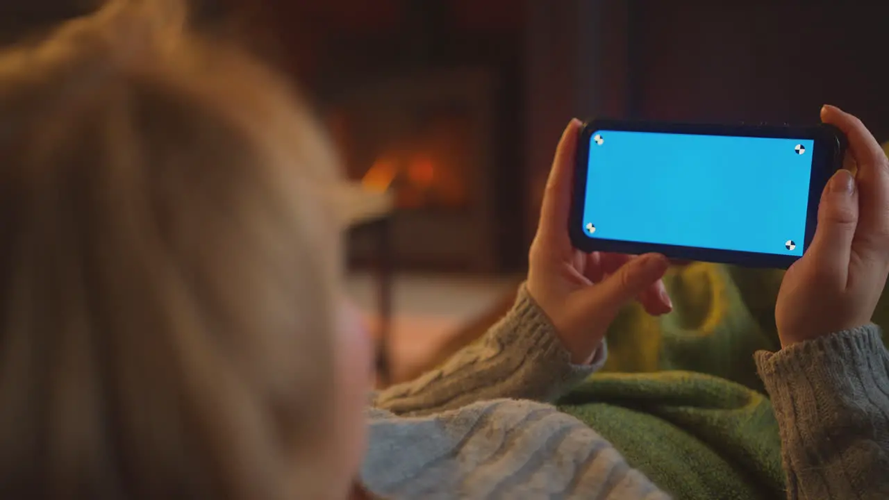 Close Up Of Woman At Home Lying In Lounge With Fire Streaming Onto Blue Screen Mobile Phone