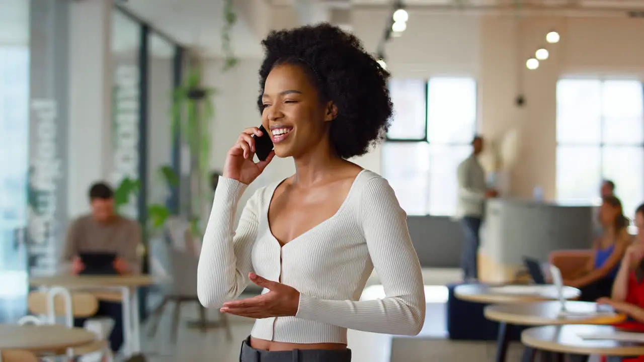 Smiling Businesswoman Standing In Busy Open Plan Office Talking On Mobile Phone