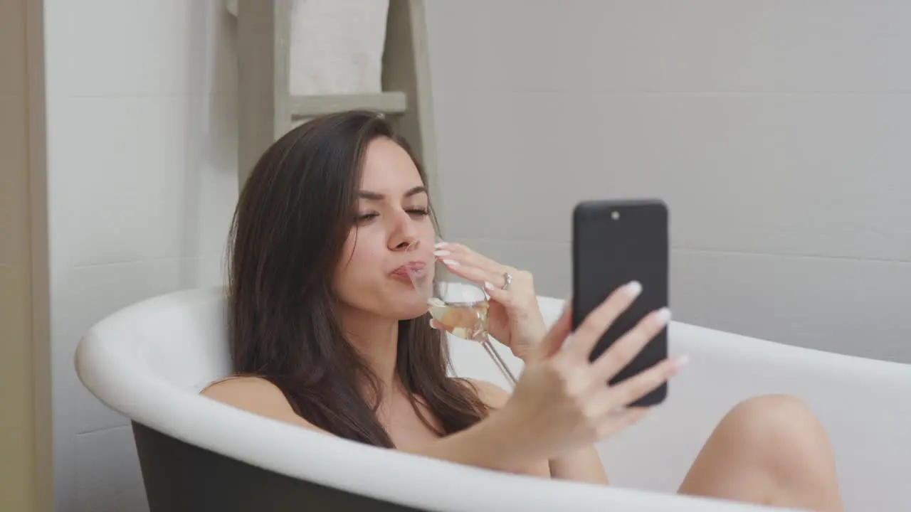 Woman Lying And Relaxing In Bath At Home Posing For Selfie On Mobile Phone And Drinking Glass Of Wine