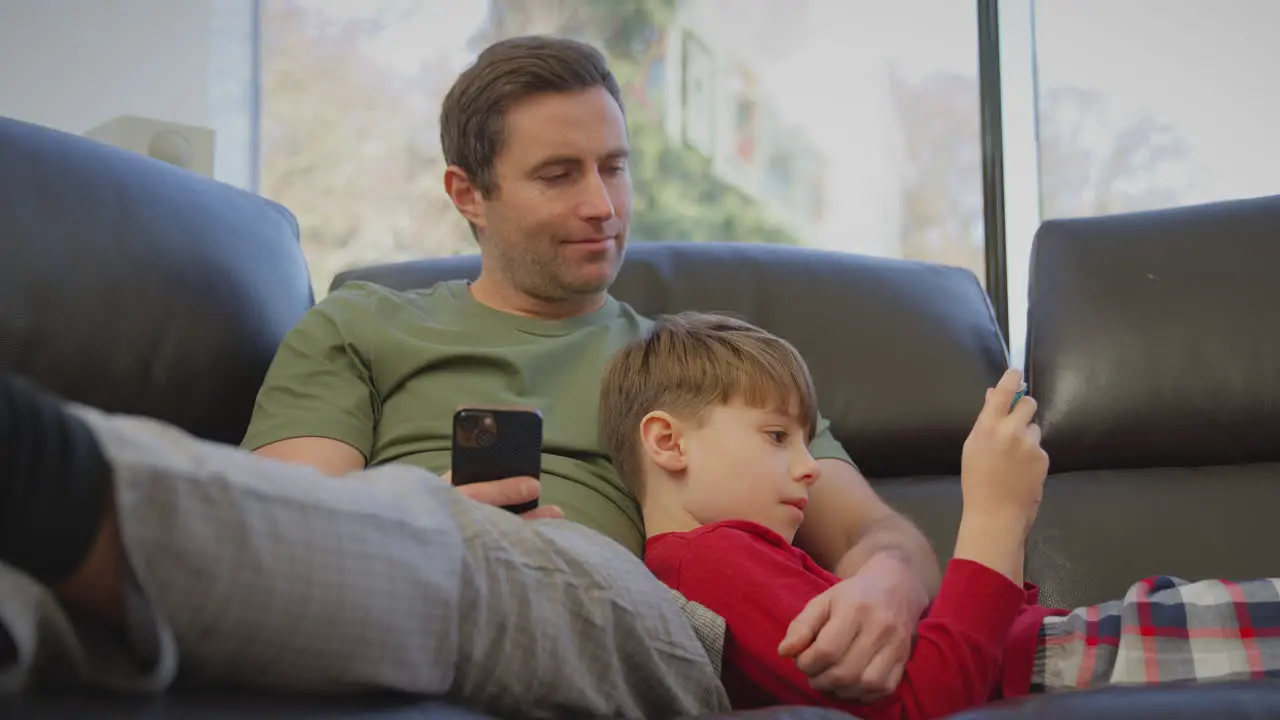 Father uses mobile phone as son plays computer game on portable gaming device lying on sofa at home in pyjamas shot in slow motion