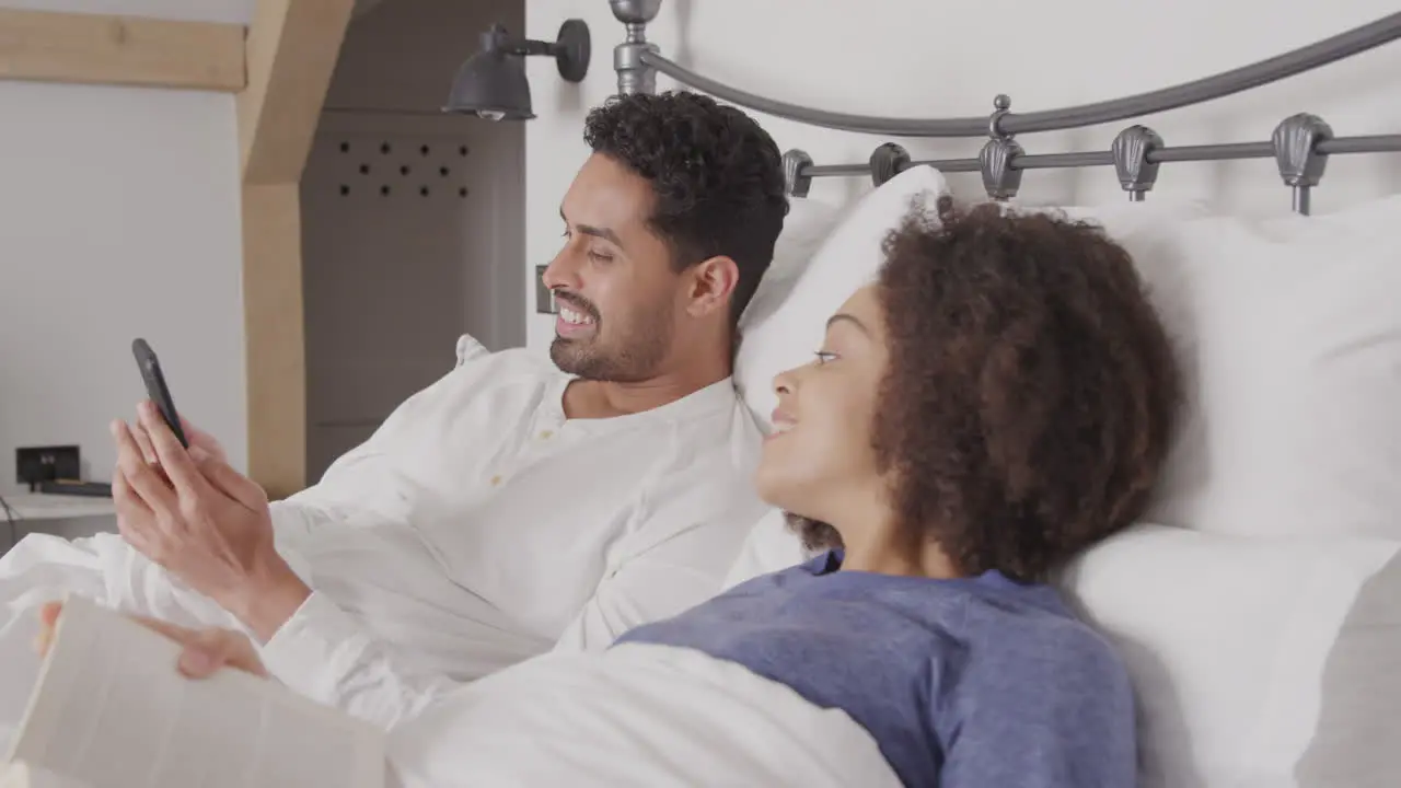 Couple In Bed Wearing Pyjamas With Woman Reading Book And Man Taking Selfie On Mobile Phone