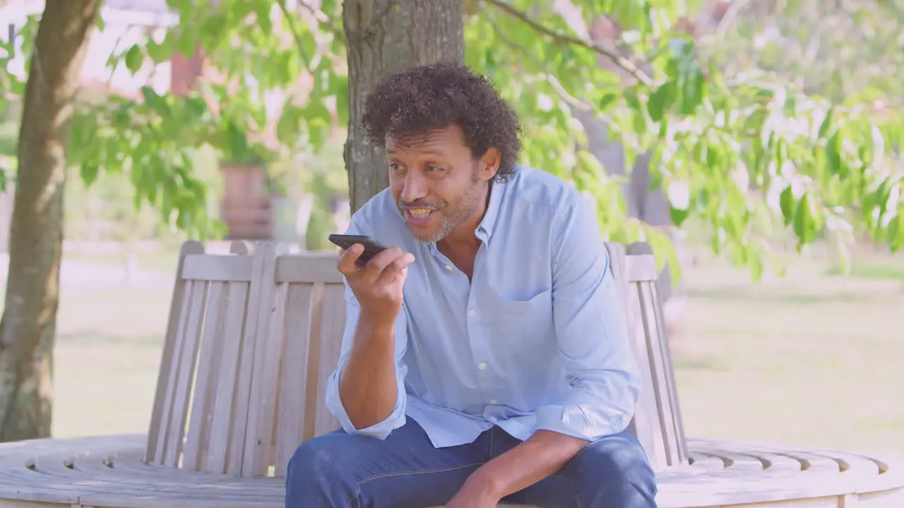 Mature Man Sitting On Park Bench Under Tree In Summer Talking On Mobile Phone Using Microphone