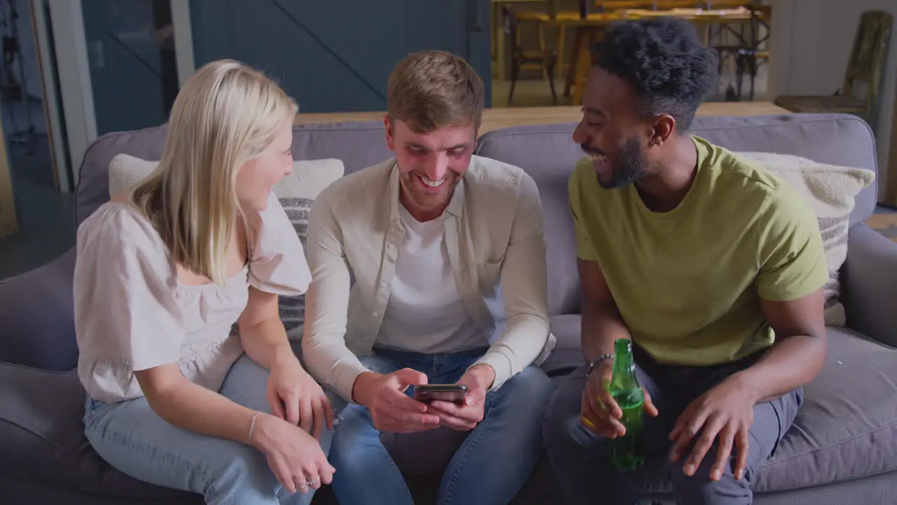 Multi-Cultural Friends Sitting On Sofa In Lounge At Home Looking At Mobile Phone Together