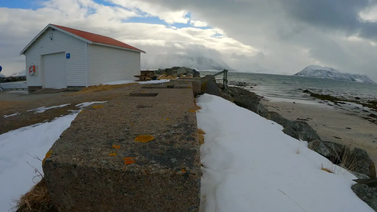 Low handheld dolly shot along breakwater wall with thick snow dark forboding clouds and mountains in winter Åse Harbor Andoya Norway