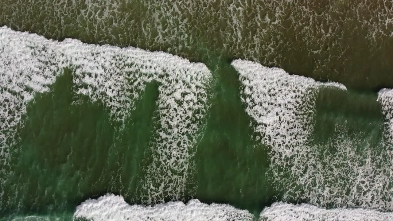 A top down drone shot of crystal clear green rolling waves heading towards the gold sands of a Cornish beach