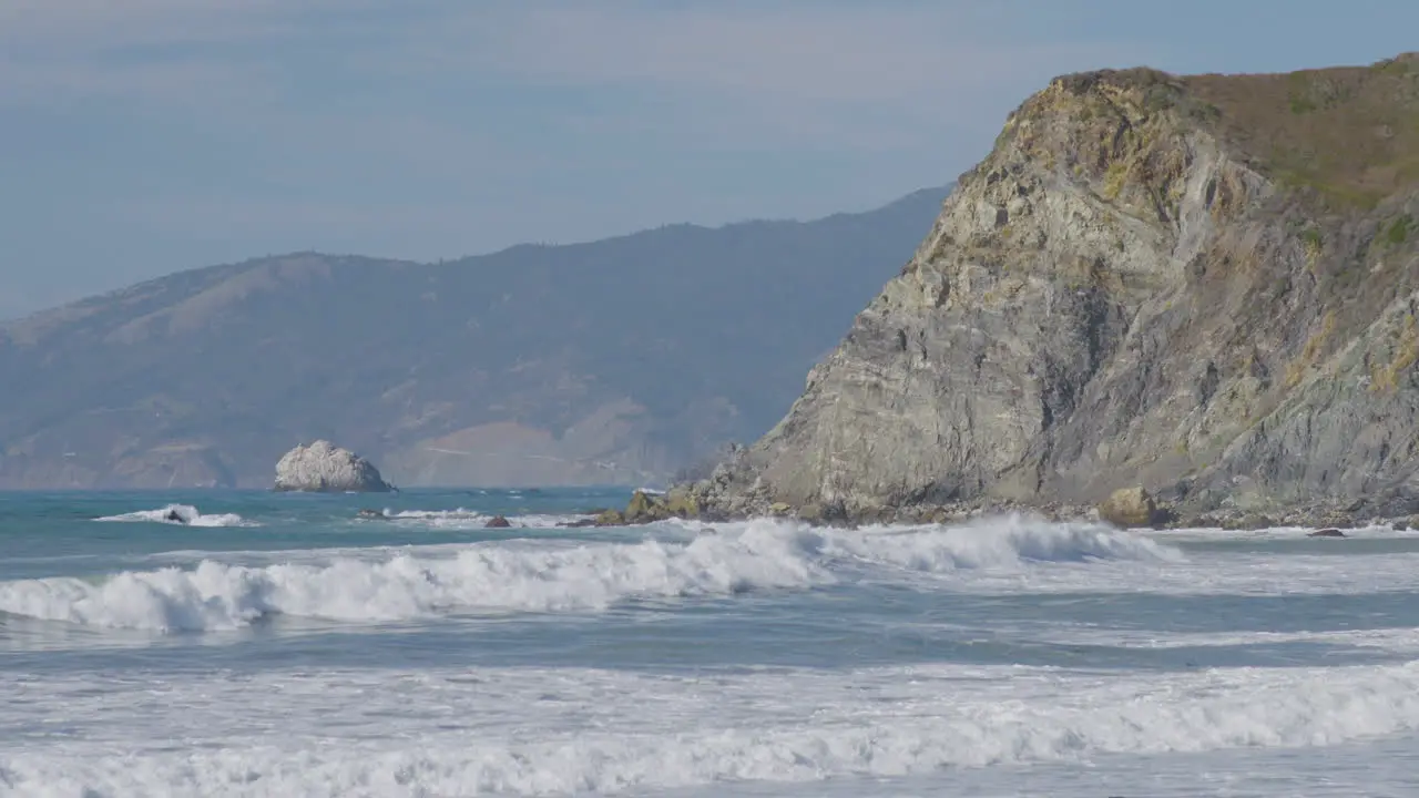 Stationary shot of waves crashing into the side of a cliff with mountains on the background located in Big Sur California Beach