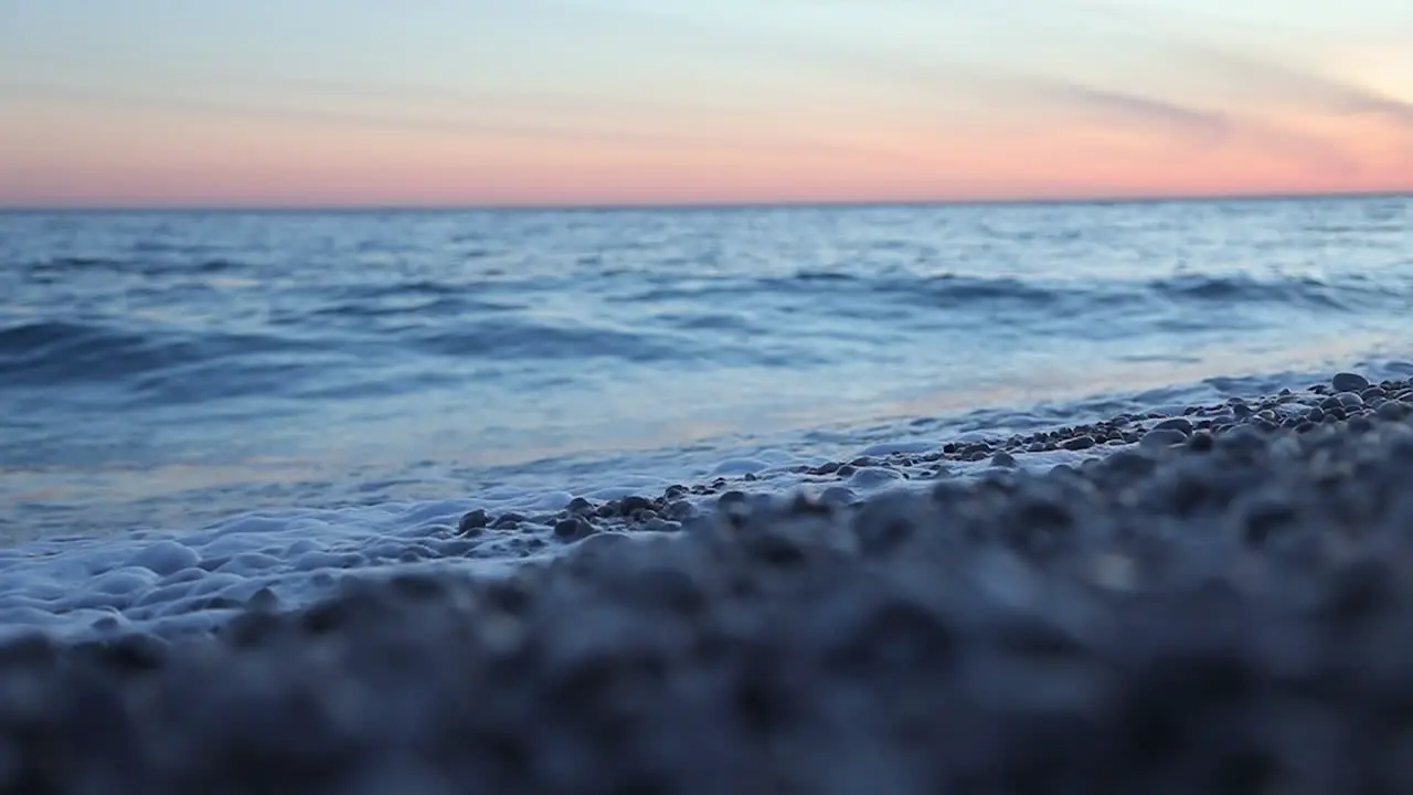 Sea waves splashing on shore with pebbles at twilight reflecting colorful sky slow motion