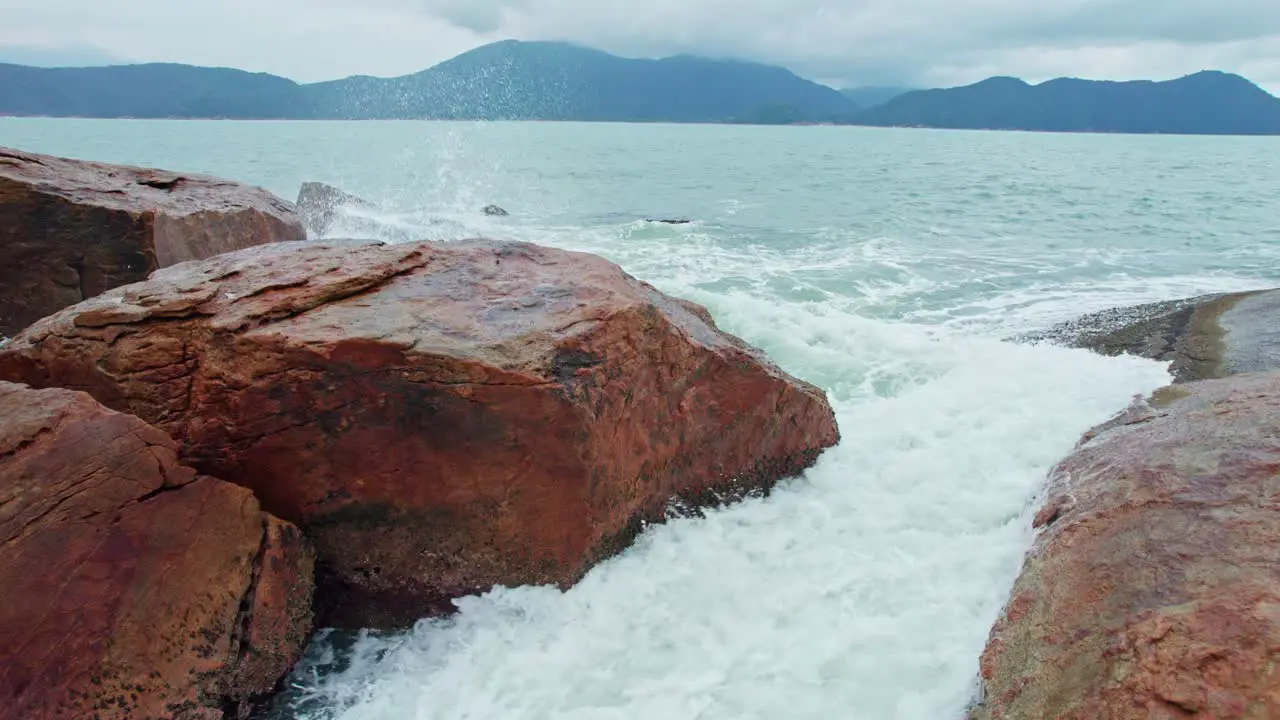 Stunning aerial forward orbital shot of green sea waves splashing on the rocks coast with white bubbles