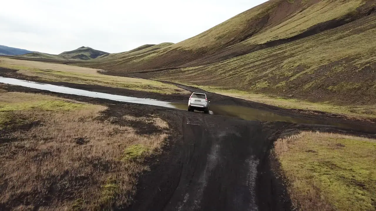 Dramatic Aerial Following Four Wheel Drive Vehicle in Iceland Landscape Crossing Stream Under Volcanic Hill