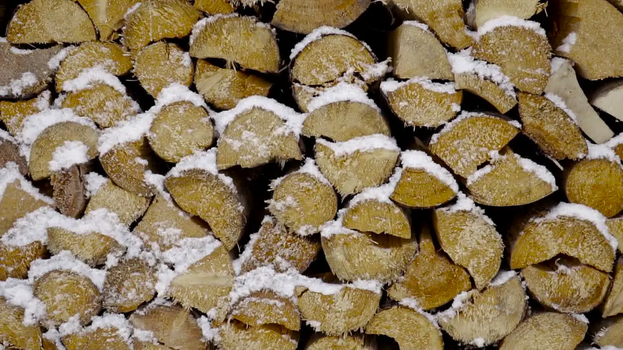 Long downward pan over cut logs of wood stacked up covered in snow with snowflakes flying through the frame