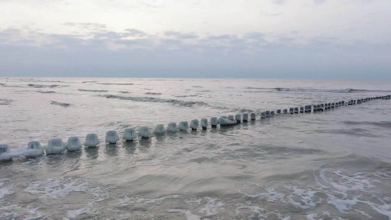 Aerial establishing view of an old wooden pier at the Baltic sea coastline overcast winter day white sand beach covered in snow ice on wood poles calm seashore wide drone shot moving forward low