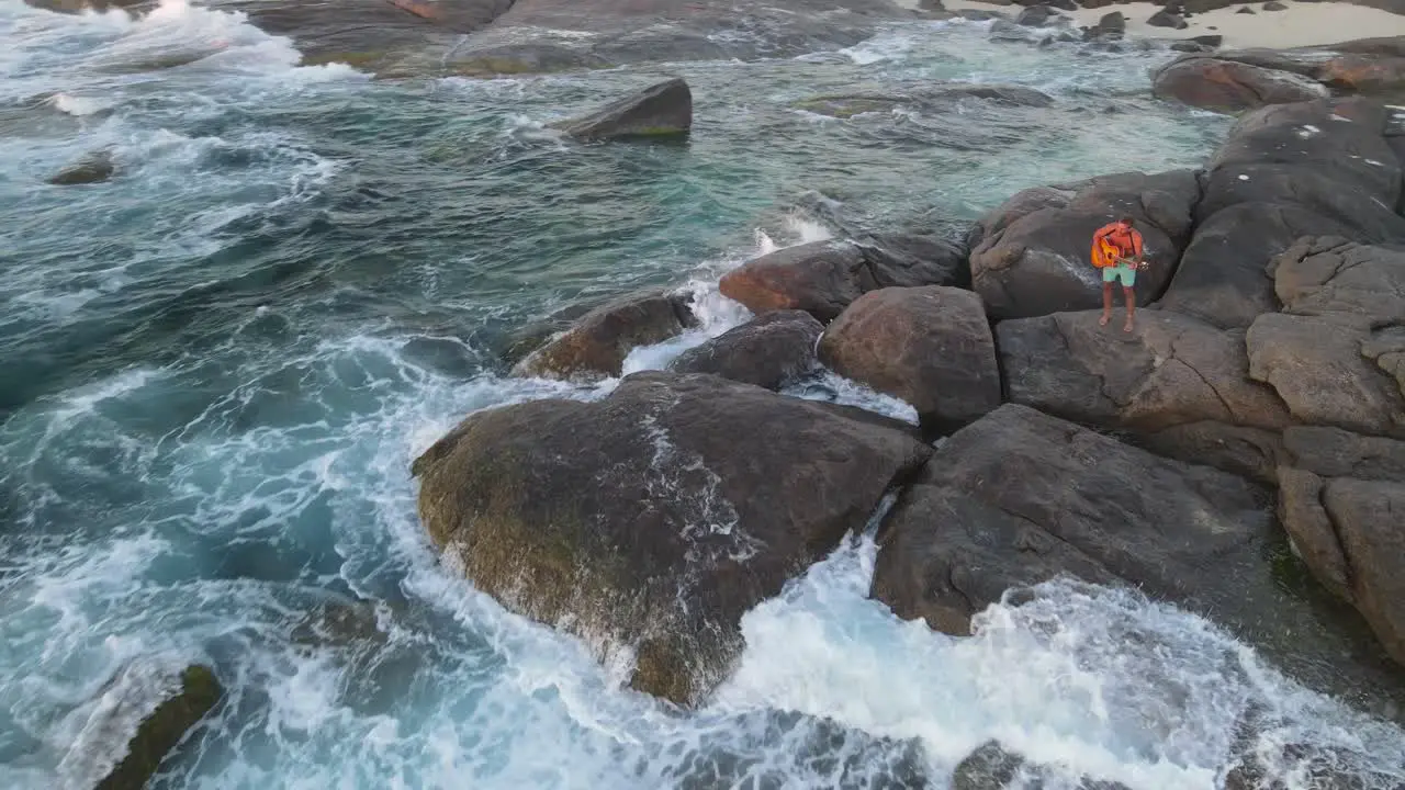 4K Amazing aerial shot attractive young male playing guitar on rocks front of wild sea waves at sunset lifestyle Slow motion 2