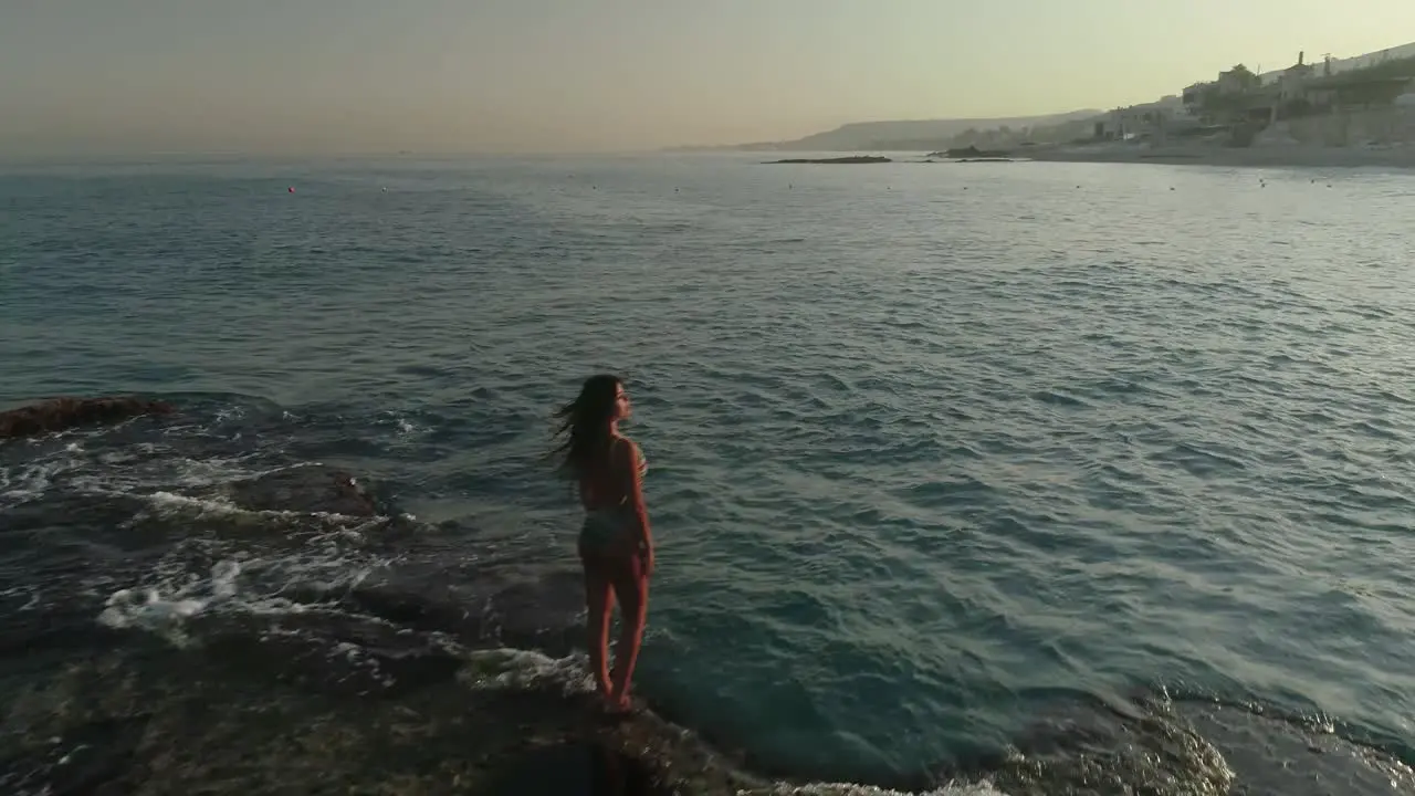 Young Caucasian woman with long brown hair wearing bathing suit stands on Batroun rock outcropping island in blue ocean sea water at sunset Lebanon overhead aerial flyover approach