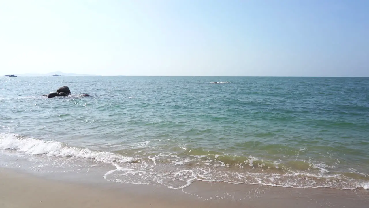 wide shot of ocean view from a tropical beach with low surf on a sunny blue sky day with rocks in the ocean and far away shoreline and islands