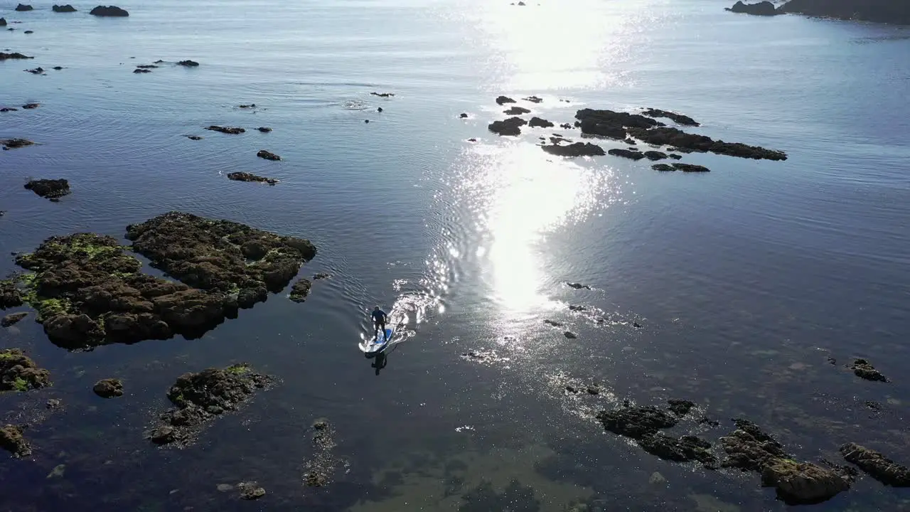 Aerial orbit of Stand up paddle boarding on clear water through rocks near a beach with sun reflecting in the background in the Atlantic