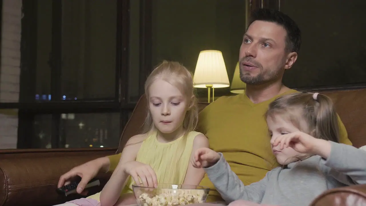 Papa Mit TV-Fernbedienung Und Seine Beiden Kleinen Töchter Essen Popcorn Und Schauen Sich Einen Film An Während Sie Nachts Zu Hause Auf Dem Sofa Sitzen 1