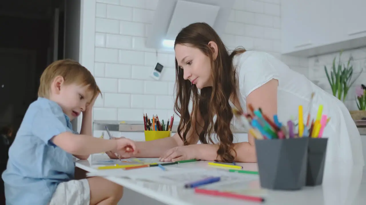 Mom helps son to perform preschool homework to draw a pencil drawing sitting at the table in the house