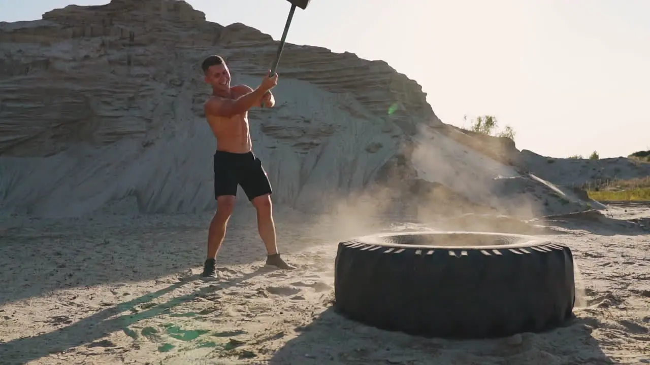 Muscle athlete strongman man hits a hammer on a huge wheel in the sandy mountains in slow motion strength and endurance training for wrestlers