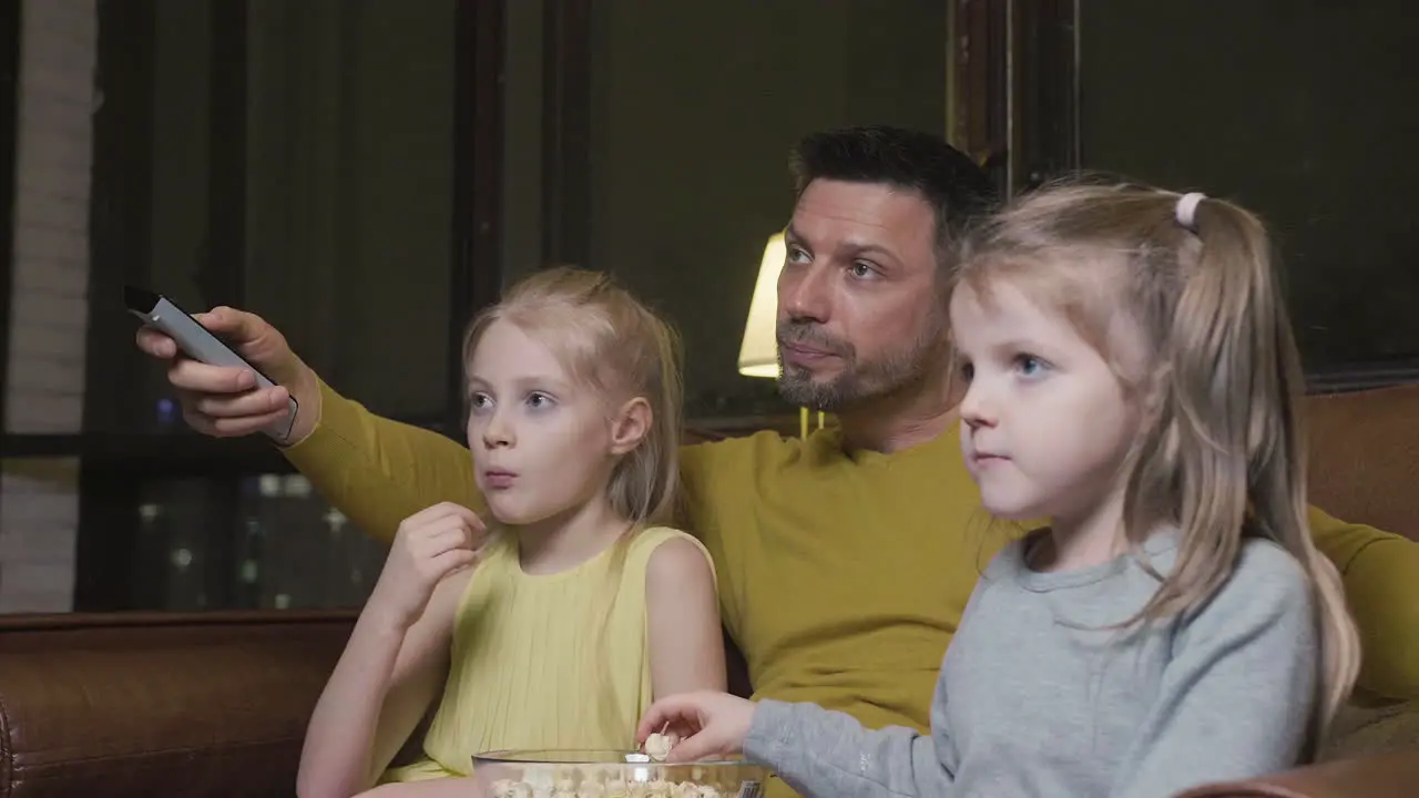 Dad With Tv Remote Control And His Two Little Daughters Eating Popcorn And Watching A Movie While Sitting On Sofa At Night At Home