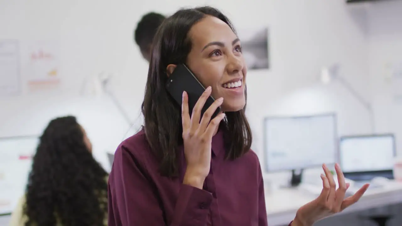 Smiling biracial businesswoman talking on smartphone with colleagues in background in modern office