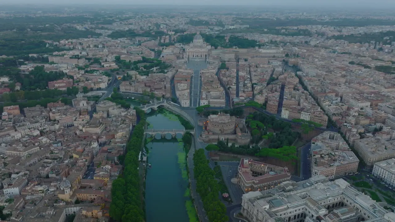 Aerial panoramic footage of Tiber river flowing through historic city centre at dusk Castel SantAngelo on riverbank and famous St Peters Basilica in Vatican City in distance Rome Italy