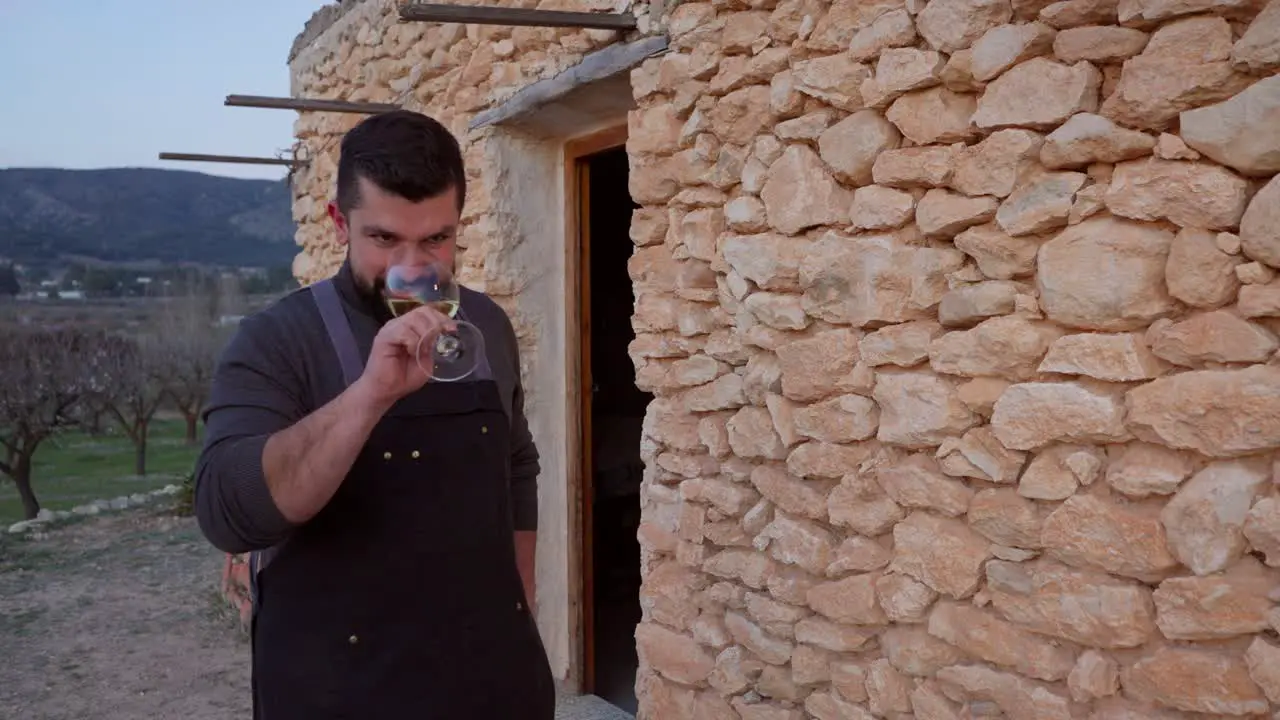 A smiling bearded man toasts with a glass of white wine in the background of a typical Mediterranean stone hut