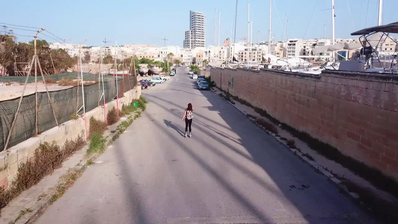 Slow mo active track drone shot of a young athletic woman inline skating on the streets from the back in the middle of parking cars waving her arms up as she is gliding through the windy breeze