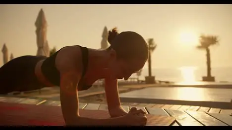 A brunette girl with starry hair in a black sports summer suit does plank exercises on a red mat on a Sunny beach at Sunrise Sunny beach closed with boards