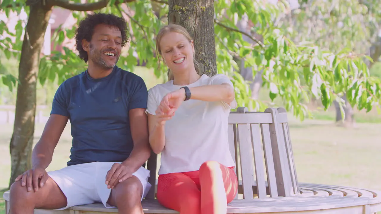 Couple Wearing Fitness Clothing Sit On Seat Under Tree Checking Activity Monitor On Smartwatch