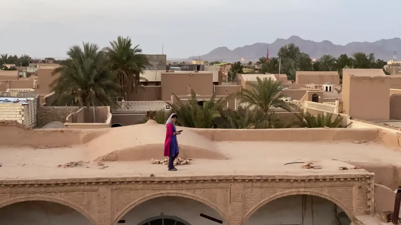 An Asian Middle East Girl Wear Scarf and Blue Galaxy Red Pomegranate Color Shirt Walking on Old Historical Ancient Club House Made by Adobe and Mudbrick by Traditional Architecture Design Iran Yazd