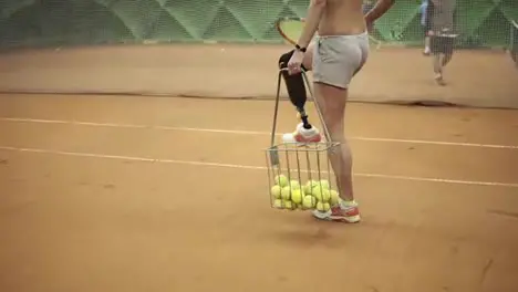 Close up footage of a young disabled girl picking up balls from a tennis court and with racket in her hand Short shorts basket for tennis balls Indoors