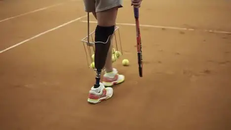 Tracking footage of a sports girl with a prosthesis on her right foot picking up tennis balls with the basket Tennis player Back view