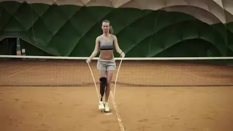 A beautiful brave girl is practicing hard with a skipping rope on a tennis court On the right foot the prosthesis is up to the knee Front view