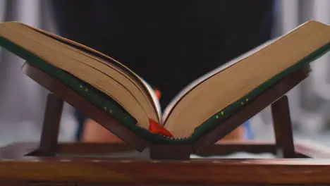 Close Up Of Open Copy Of The Quran On Stand At Home With Woman Sitting Behind