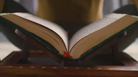 Close Up Of Open Copy Of The Quran On Stand At Home With Man Sitting Behind