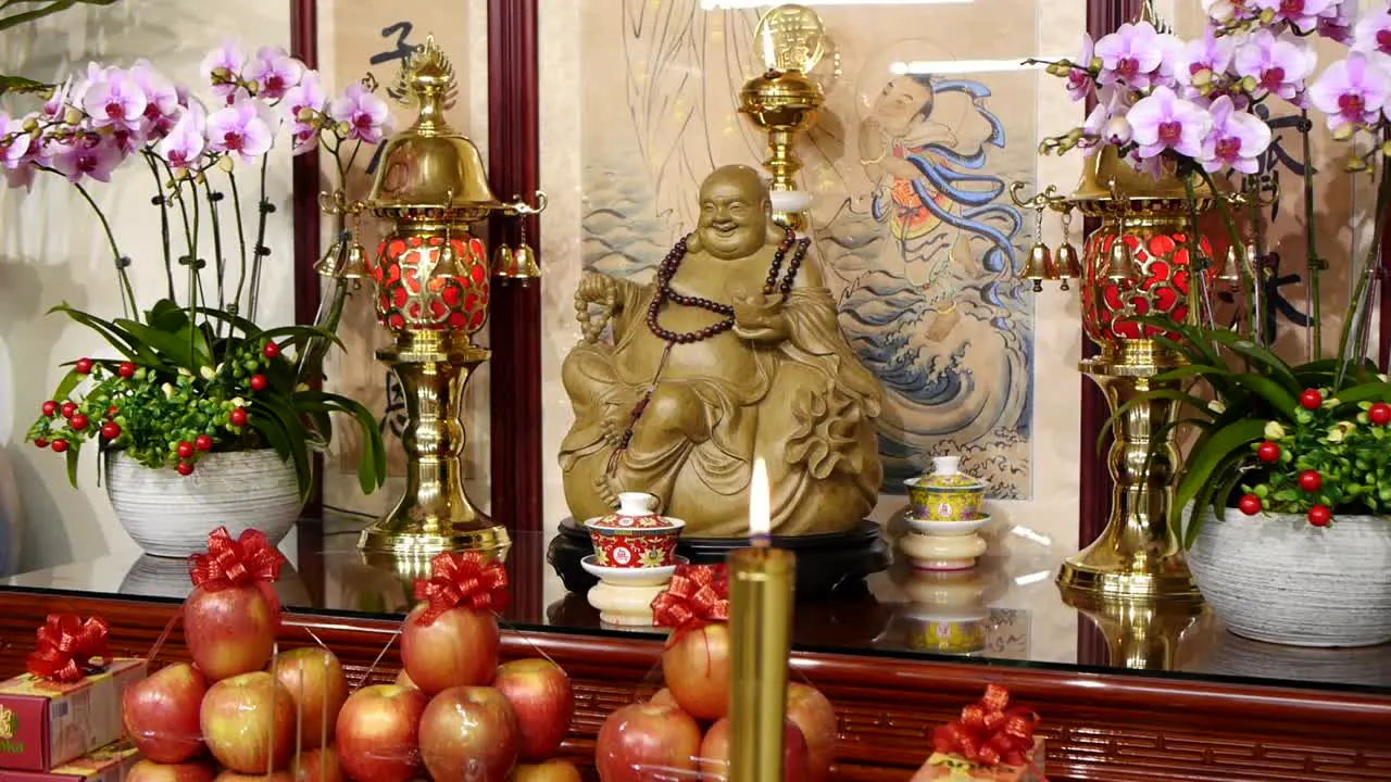 Carved wooden laughing Buddha sitting on shrine with fruits and candles decorated in front with camera moving downward