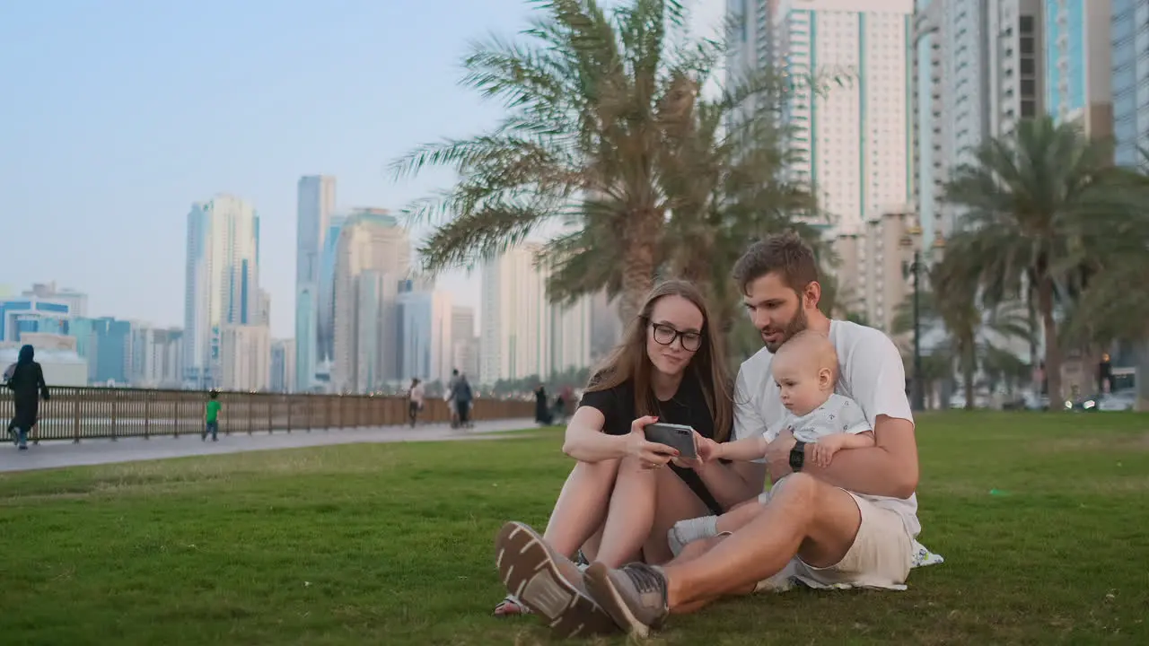 Happy family with one children sitting together on grass in park and taking a selfie With smartphone