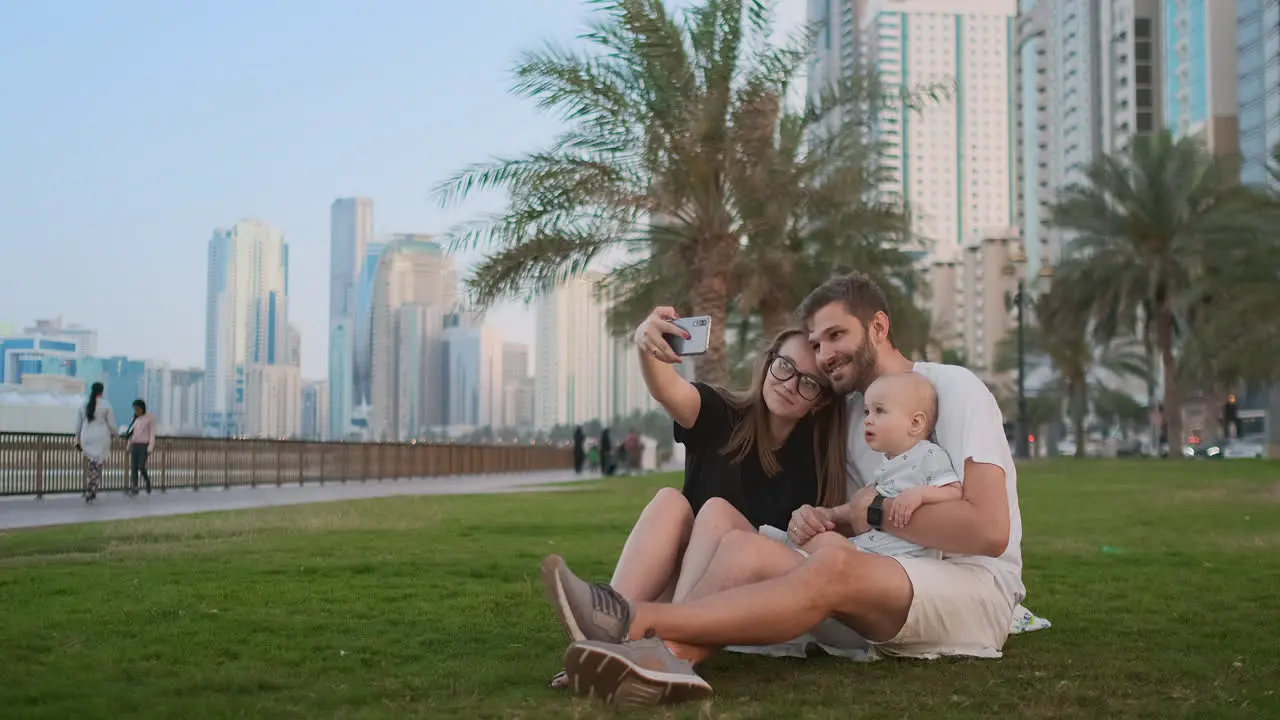 Happy family with two children sitting together on grass in park and taking a selfie With smartphone