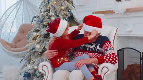 Grandchild girl kid fixes Christmas Santa Claus hat on happy senior old smiling grandfather at home