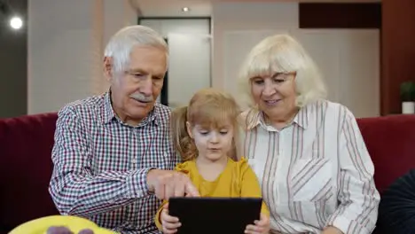 Child girl shows something in laptop to grandparents seniors couple learning how to use tablet