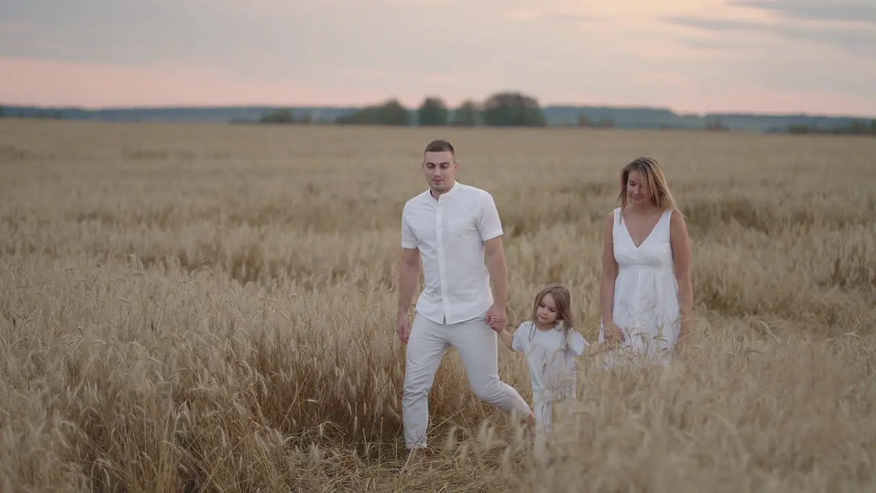 Young couple of parents with girl children holding hands of each other and running through wheat field at sunset Happy family jogging among barley meadow and enjoying nature together Slow motion